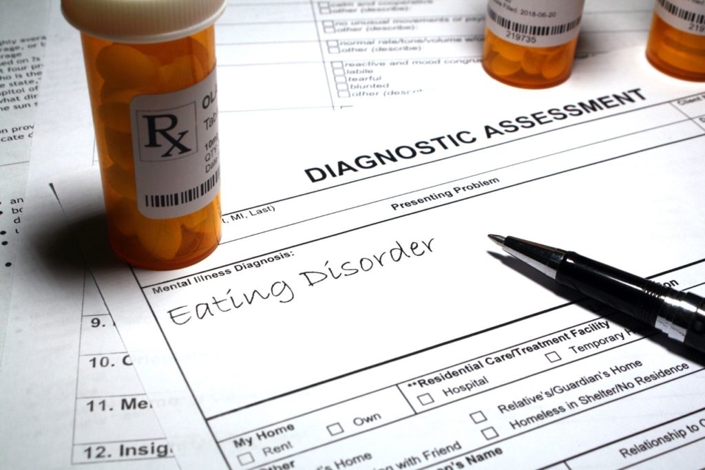 Image of a doctor's diagnostic assessment sheet. The sheet says Eating Disorder as the mental illness diagnoses. On top of the paper sits orange medication bottles with white pills.