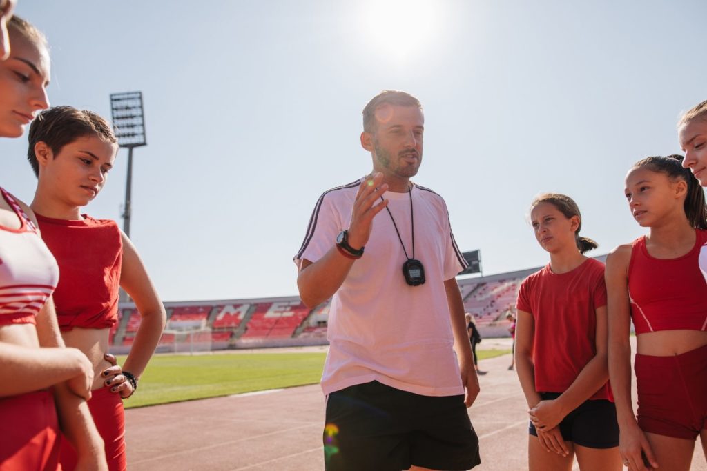 Image of a male coach talking to his team of young female athletes. The coach wears a white top and a stopwatch around his neck. The athletes wear red uniforms as they listen intently.
