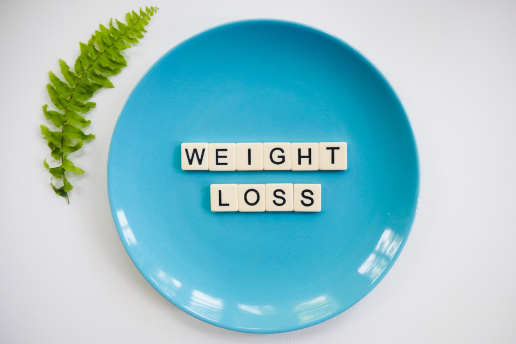Birds eye view of a robin egg blue plate on a white surface. On the plate is scrabble letters that spell "weight loss" to represent diet culture and dieting. To the left of the plate is a single green, fern leaf.
