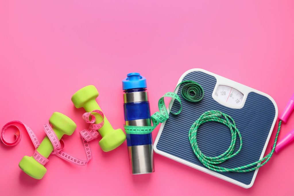 Overhead view of typical symbols used to represent weight loss. On a pink background sits a bathroom scale, a jump rope. a water bottle, and a set of dumbells. The water bottle and dumbbells are each wrapped in their own measuring tape.