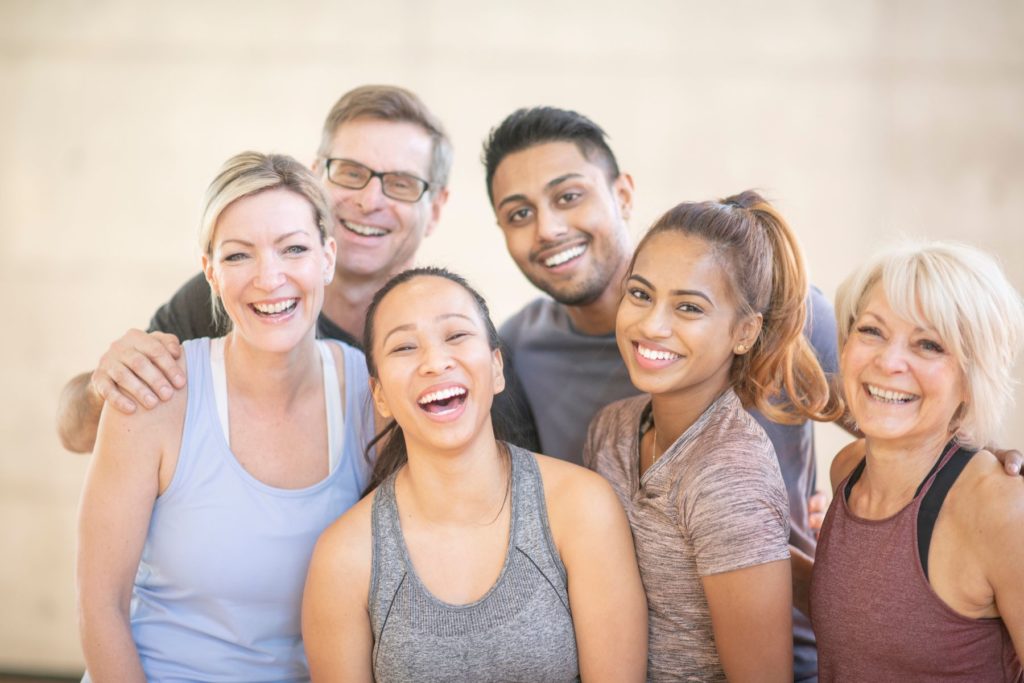 A group of 4 women stand in front of two men as they huddle together to pose for a photo. The group is all dressed in athletic wear as if they just completed a workout class together. The group appears to be smiling and laughing as they look at the camera.