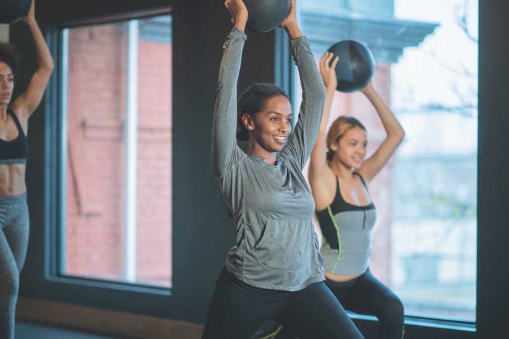Image of three women in an exercise class. All of the women are smiling as they hold a weighted exercise ball in their hands as they hold a lunge position.