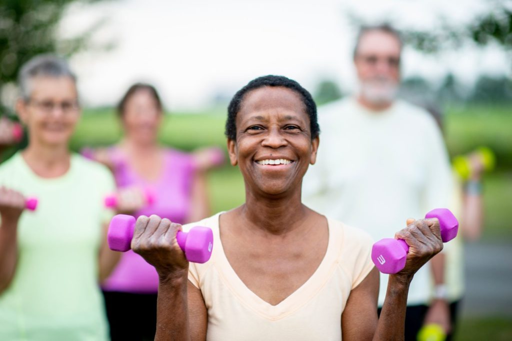 Image of a group of older adults doing an exercise class. The woman in the front of the image is in focus while the people behind her are out of focus.  The woman in the foreground holds a magenta three pound dumbbell in each hand as if she were doing a curl as she looks at the camera smiling. 