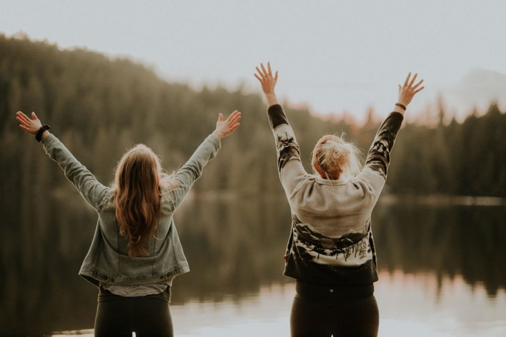 Two women stand with their backs to the camera. They are facing a lake that is surrounded by evergreen trees. The two women have their hands up in the air as they express joyful movement.