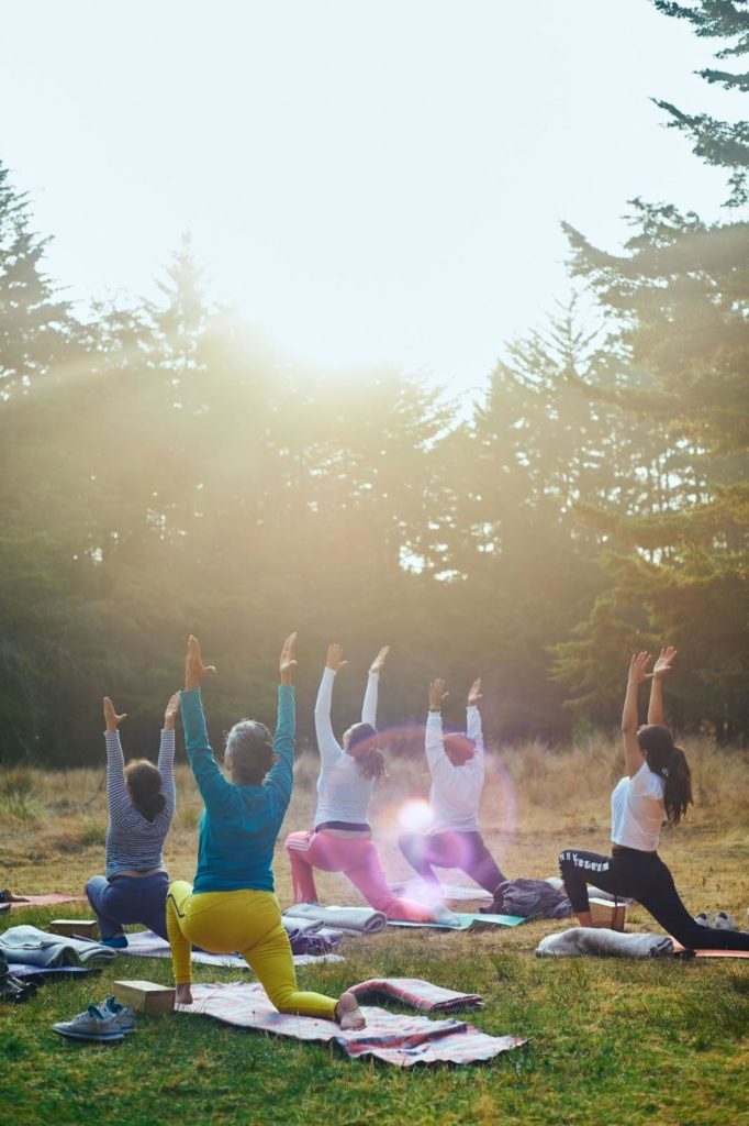 An image of a group of women performing yoga outside. The women have their yoga mats rolled out on the grass. The women are holding a lunge with their back knees on the ground and hands in the air above their heads. In the background is a row of evergreen trees. 