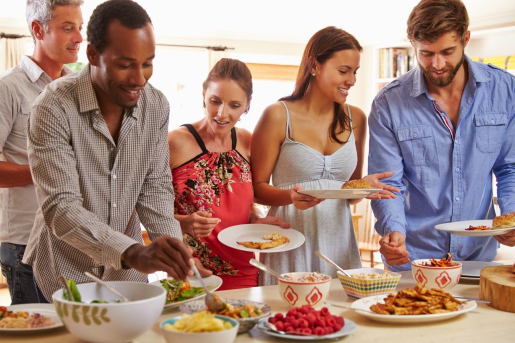 Five friends standing at a kitchen counter holding plates of food. The counter is covered in a spread of various foods as the friends serve themselves from the dishes.