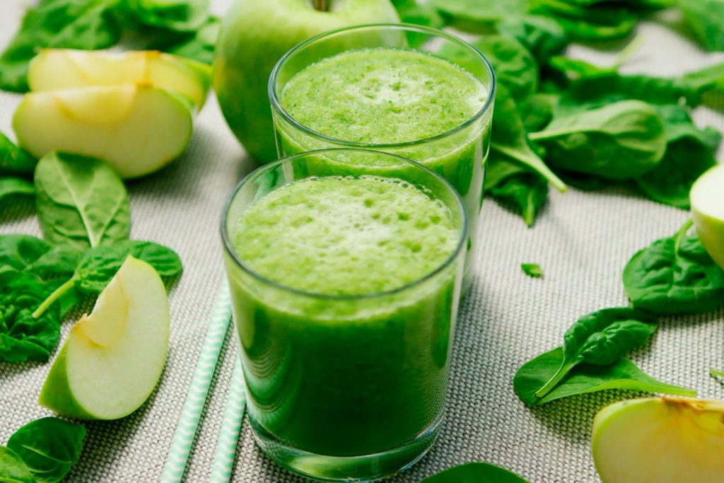 Two cups of green juice on a burlap tablecloth. Sprinkled around the cups of juice are spinach leaves and slices of green apple.