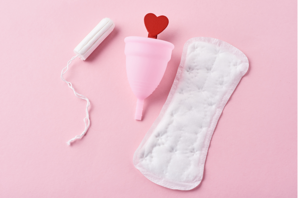Overhead view of a layout of women's feminine or period products. On top of a light pink background lays a light pink menstrual cup in the centre, a white tampon to its left, and a white panty liner to its right. There is a red heart coming out of the top of the menstrual cup.