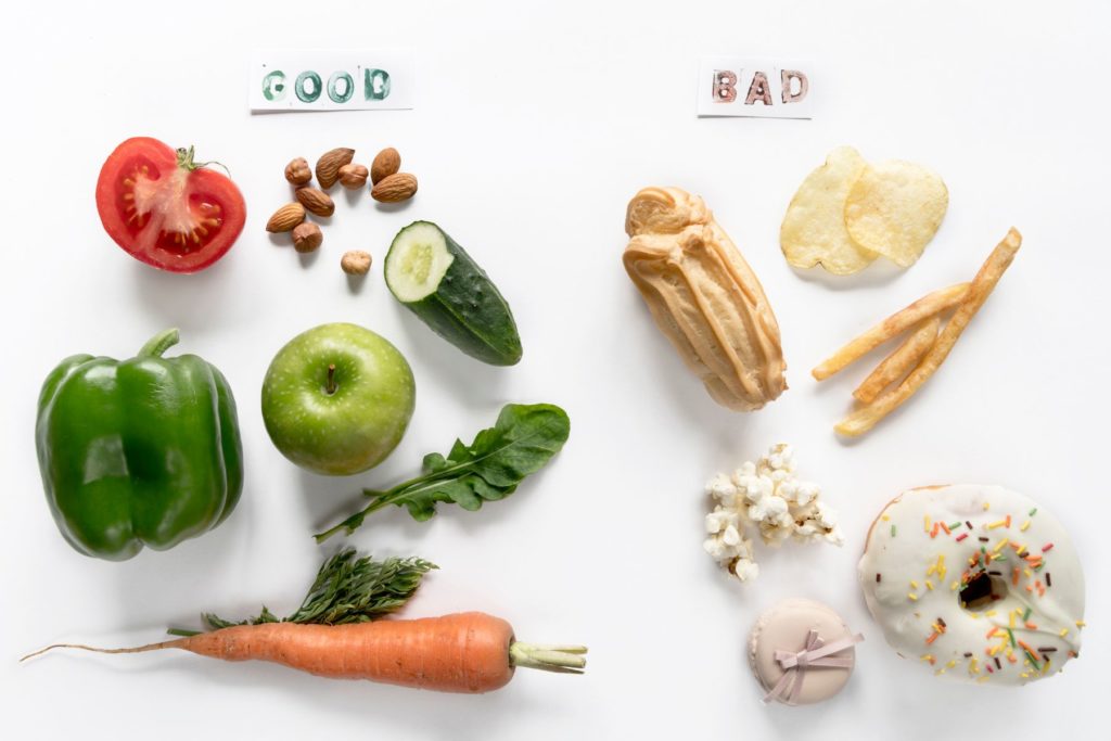 Foods laid out on a white background and grouped into two groups. One group includes fruits, veggies and nuts with a label above that says good. The other group includes doughnuts, fries, chips and other snack foods and has a label above that reads bad.