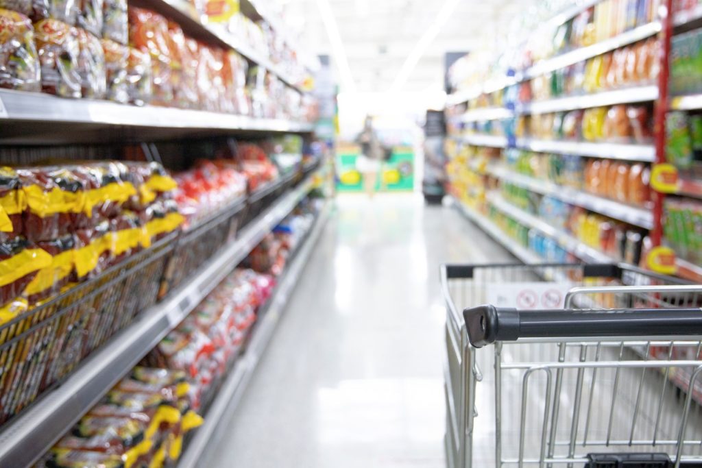 A grocery cart is in focus in the right hand side of the image. On either side of the image are the aisles of a grocery store that blur into the distance. The aisle contains snack foods like chips and cookies.