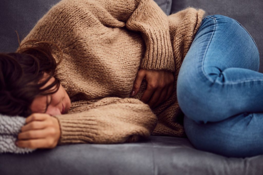 Image of a woman curled up in the fetal position on a couch holding her stomach. The woman's face appears to be in pain. The woman wears blue jeans, a tan sweater, and hea dark brown hair.