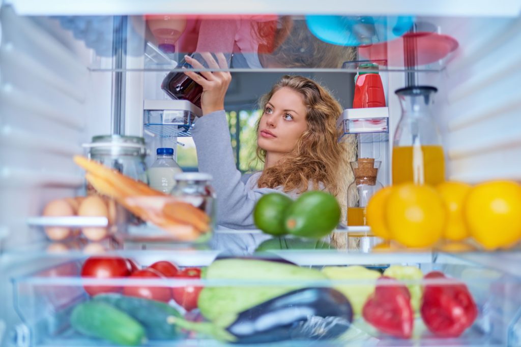 Image taken as if we are inside of a fridge looking out at the woman who is picking out what she is eating. The woman has blond, curl hair and is pulling a jar out of the fridge. The fridge is filled with colourful produce, some eggs, and some glass containers of juice.