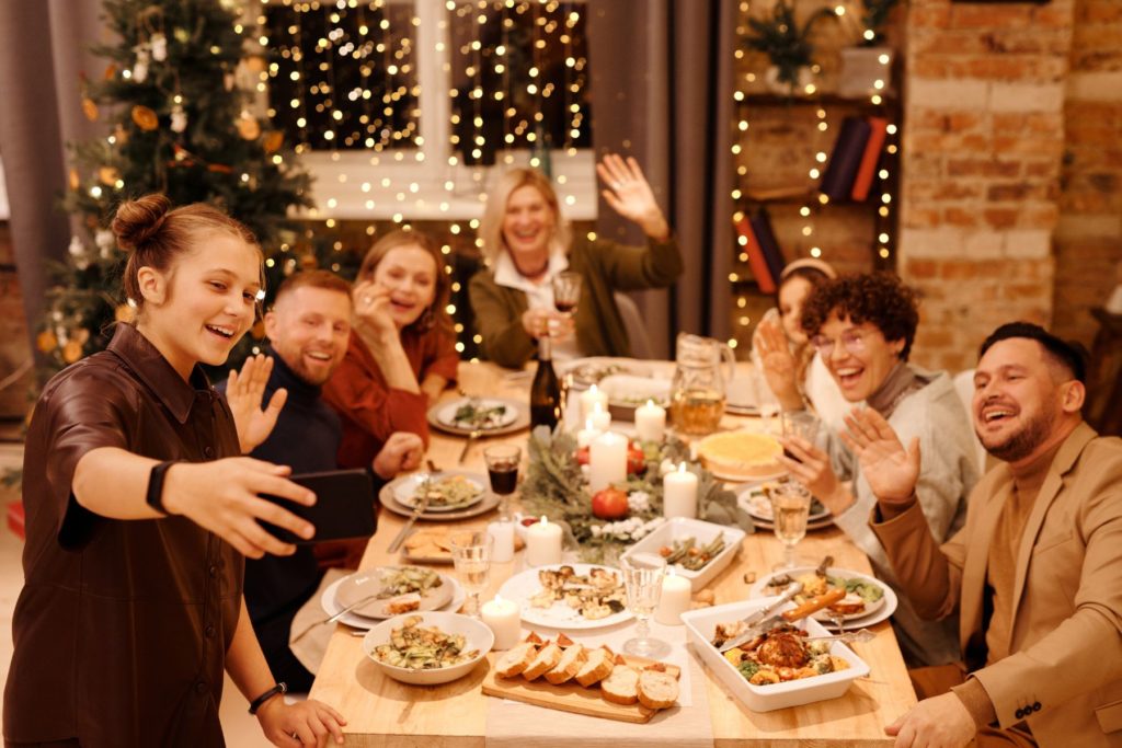 Image of a family sitting around a long table for holiday supper. a young girl holds up a phone as if she is facetiming someone. The family sitting at the table is smiling and waving at the phone. 