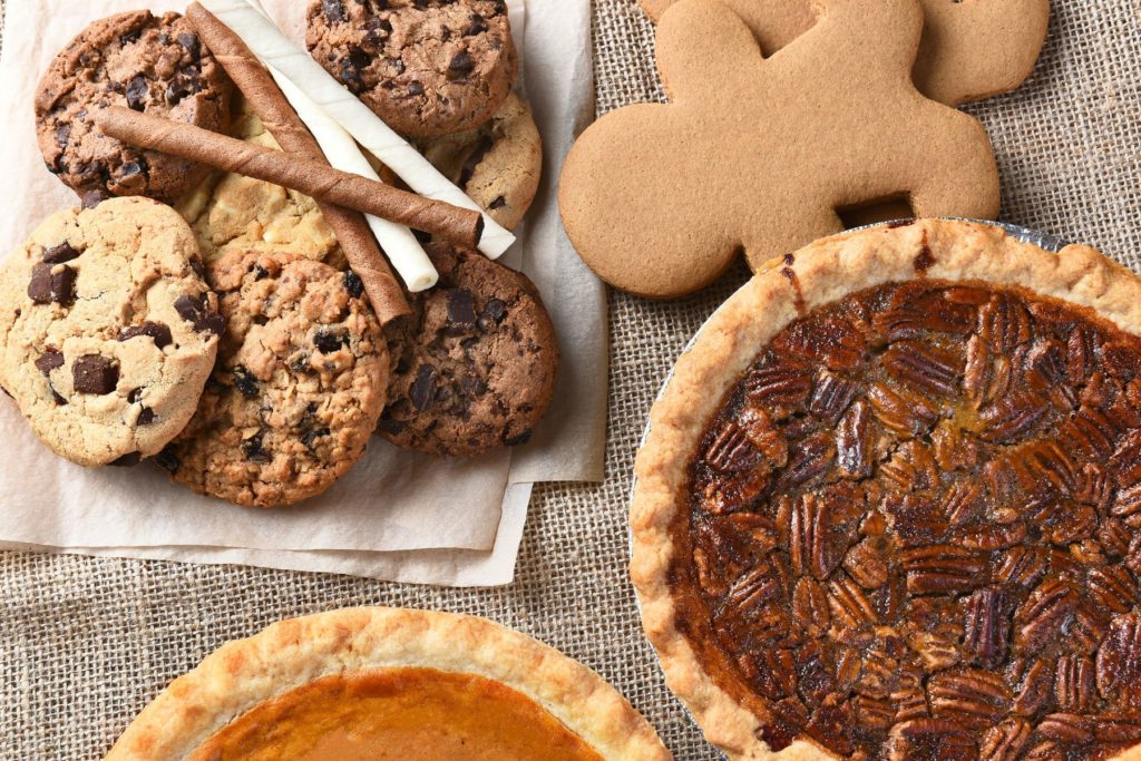 Spread of various holiday desserts. THe top right corner has chocolate chip cookies and the top right corner has plain gingerbread men. On the bottom of the image is a pumpkin pie and a pecan pie. 