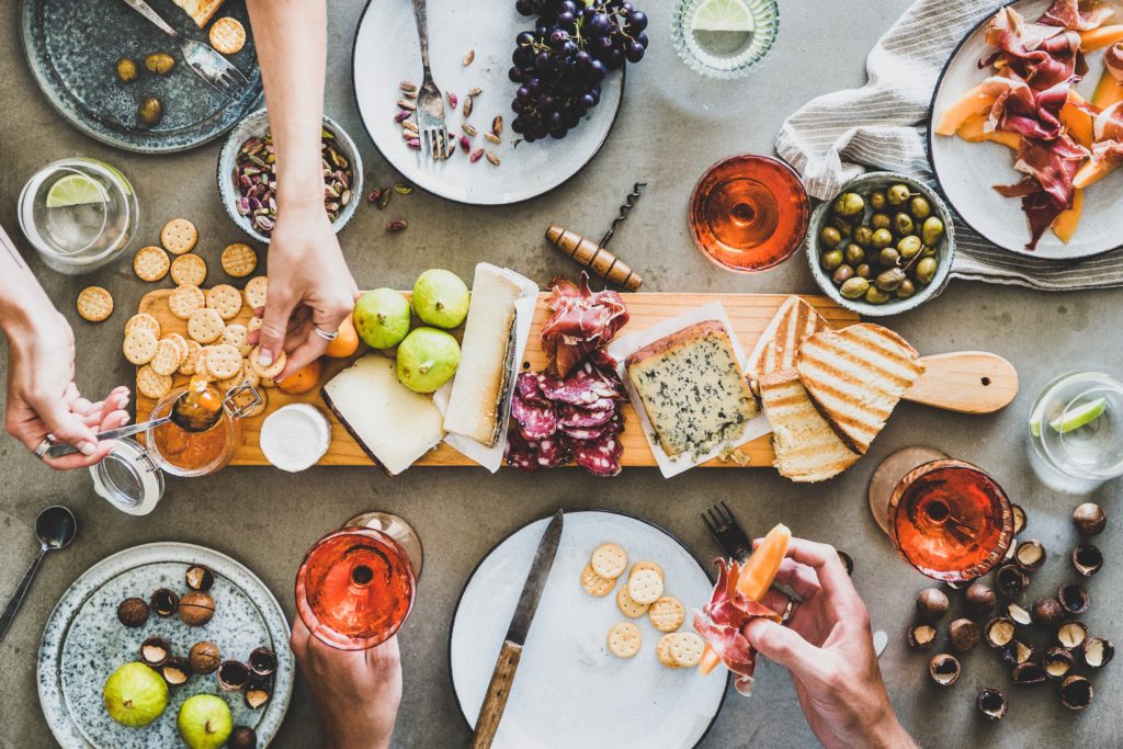 Birds Eye view of a charcuterie board, possibly served during the holidays. The board is covered in cheeses, meats, crackers, and a spread. Around the board are plates and glasses of rose. Two sets of hands reach into the frame to take food off of the board.