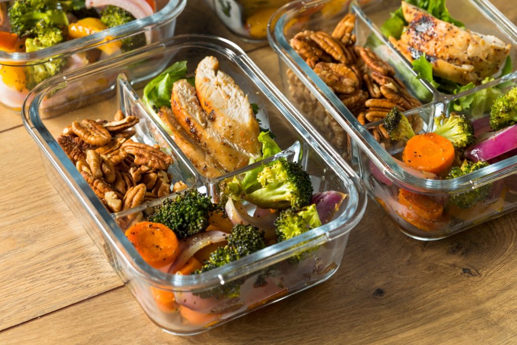Several divided glass tupperware containers hold  chicken, pecans, and roasted veggies, possibly leftovers from the holidays. The containers sit without lids on a wooden table surface.