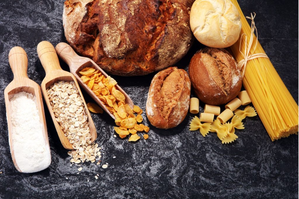 An assortment of carbohydrate containing foods are displayed on a black marble surface. Three wooden scoops sit on the left hand side. One is filled with white flour, one with oatmeal, and the other with corn flakes. In the centre sits on large, round loaf of crusty bread and three smaller dinner rolls. On the left is a bundle of raw spaghetti tied with a string with several pieces of bowtie and ditalini pasta displayed beside it.