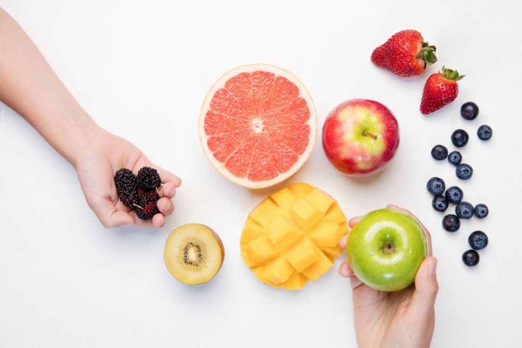 A variety of assorted fruit is assorted on a white background to represent health eating habits. The spread includes half of a grapefruit, half of a mango. a red apple, half of a yellow kiwi, 2 strawberries, and blueberries. An arm enters from the left side of the image holding blackberries in its palm. A second hand comes from the bottom right side of the image holding a green apple. 