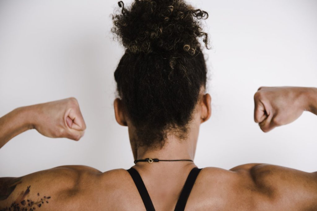 Image taken of a muscular woman's upper back. The woman has darker skin and dark curly hair. She holds her arms up as she flexes her biceps.