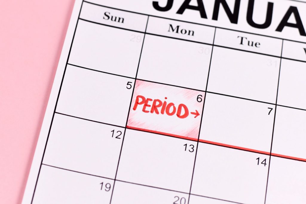 Part of a white calendar fills the frame on a pink background. The calendar reads January. On the sixth day of the calendar the word Period is written in red marker. 