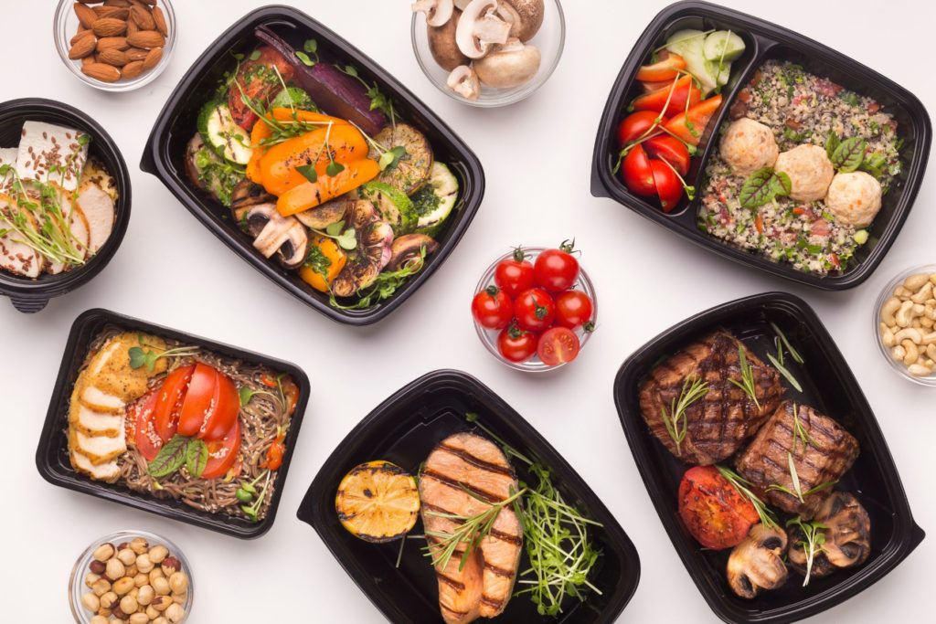 Birds-eye view of several, black meal prep containers. The containers each have a different, healthy meal. between the containers are small glass bowls filled with different ingredients such as tomatoes, mushrooms, and nuts. 