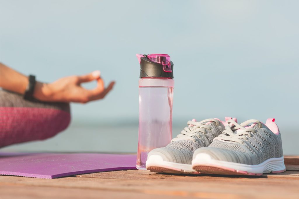 A translucent, pink water bottle sits on the ground to the left of a pair of grey and pink running shoes. On the left hand side of the image is the hand and knee of a woman just out of the frame who appears to be seated cross legged to meditate. The woman sits on a pink yoga mat. 