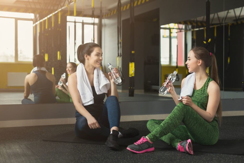 Two young women sitting on the floor of a gym facing each other. Behind the girls is a large gym mirror that covers the wall. In the mirrors reflection we can see multiple TRXs hanging from the 
ceiling. The women are drinking bottles of water rather than sports drink as they smile and laugh. 