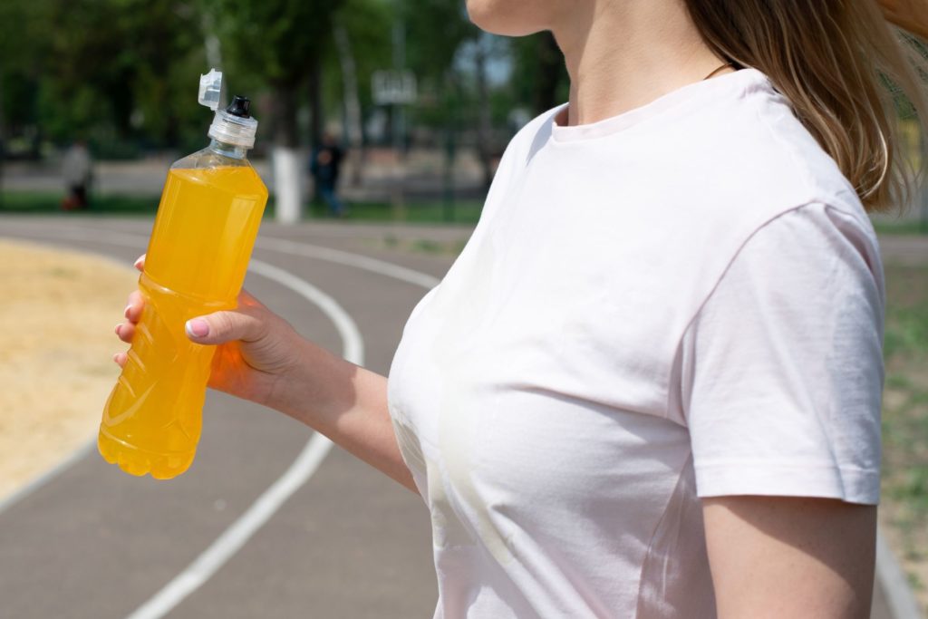A woman in a white shirt holds a clear bottle of orange sports drink. The woman appears to be bringing the bottle up to her mouth to get her hydration in. She is outside on a grey running track.