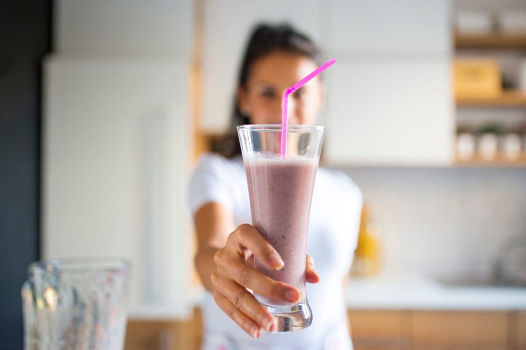 Image of a woman with dark hair in a ponytail and a white t-shirt standing in a kitchen. The woman has her arm reached out towards the camera holding a pink smoothie in a clear glass with a pink, plastic straw. The woman is out of focus while the smoothie is in focus. 