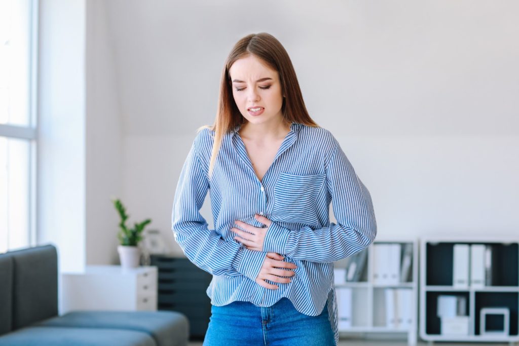 The waist up of a woman stands in the middle of the image. The woman appears to be in a living room that has a grey couch to her left and short, white bookshelves behind her. The woman wears a blue striped blouse and blue jeans. She holds her stomach as she sumpls over slightly. The woman is grimacing as if she is in pain, possibly due to IBS or her eating habits. 