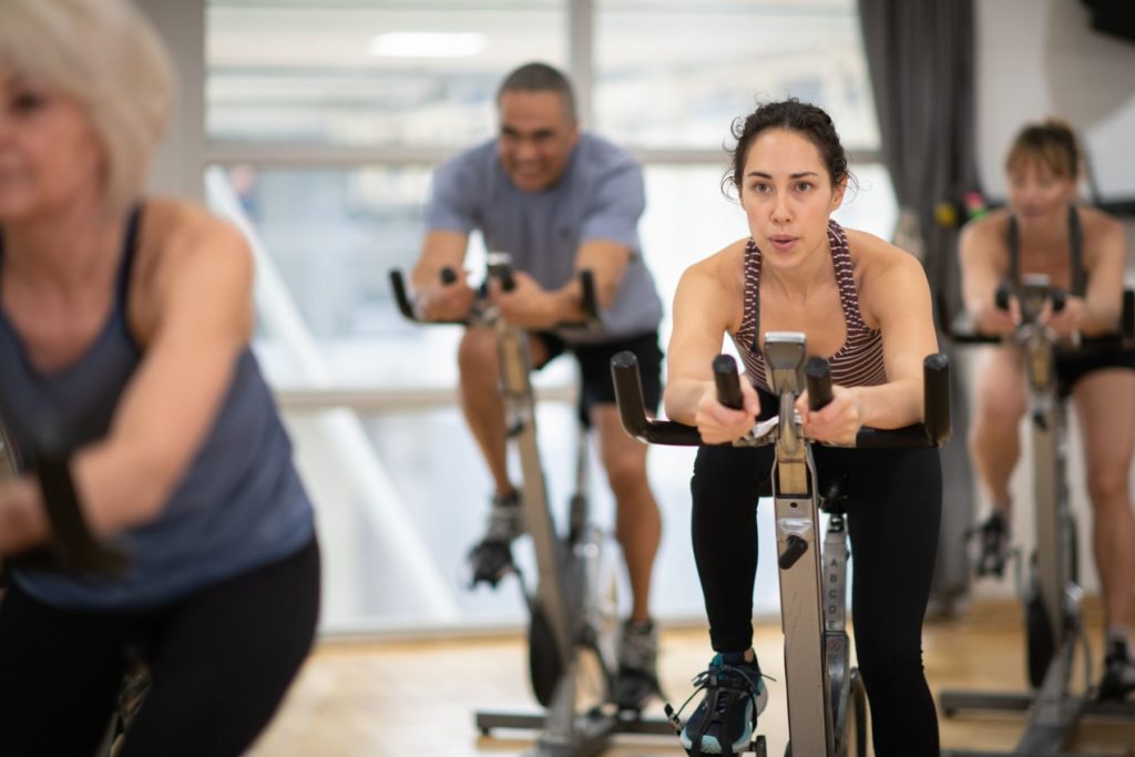 Woman with brown hair in a ponytail is sitting on a spin bike. The woman appears to be in a spin class. Two women and one man are around her, out of focus, sitting on spin bikes. The woman has her lips pursed as if she is focusing on breathing out as she completes her workouts.