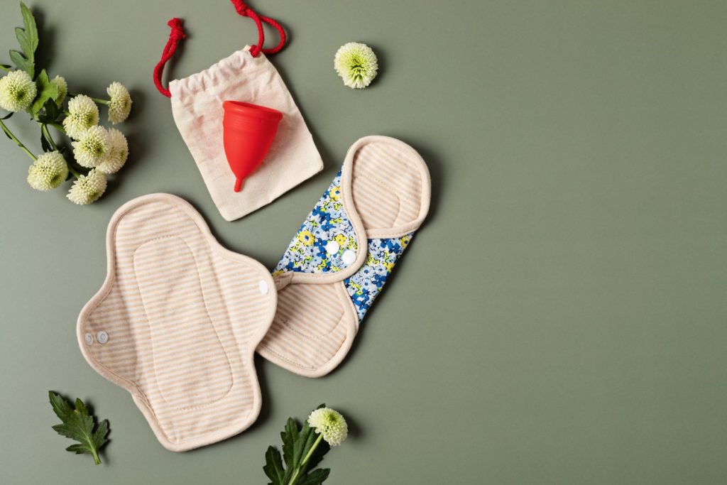 Birds Eye view of a spread of reusable products for periods on a sage green backdrop. On the left side of the image there lay two cloth menstrual pads, one red menstrual cup, and white flowers arranged around the products. 