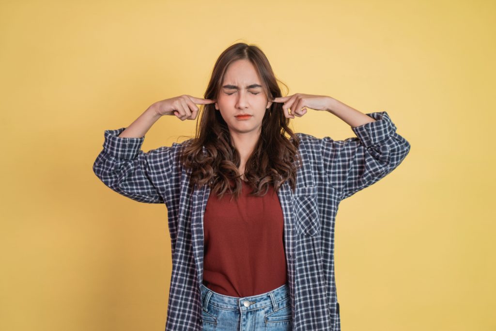 Image of a young woman standing in front of a yellow background with her fingers in her ears and her eyes closed as if it were too loud. The woman wears jeans, a red tshirt, and a black and white flannel button down open overtop. SHe has dark brown hair that is curly. 