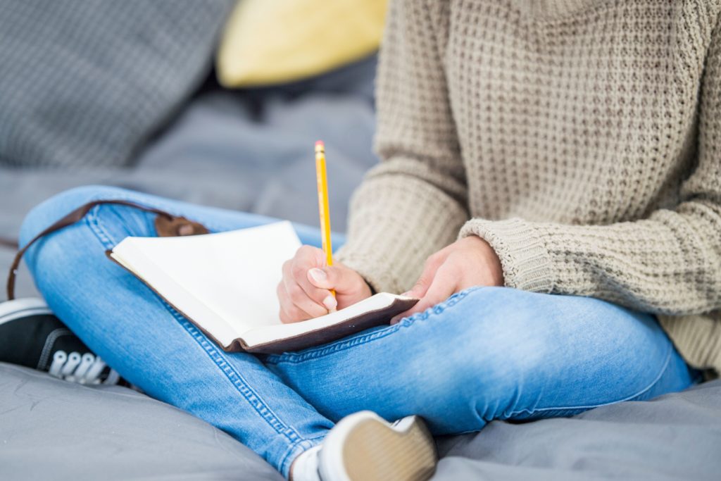 The torso and legs of a woman sitting on a grey couch or bed. The woman wears a beige knit sweater, blue jeans, and black vans. Her legs are crossed as she writes with a yellow pencil in a brown leather journal.