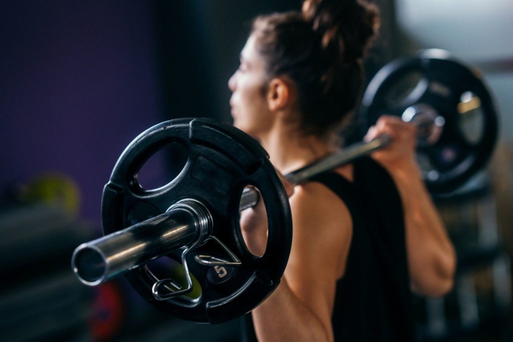 A woman in a black tank top with dark hair in a bun stands facing away from the camera. She has a barbell with black weights on her shoulders as if she is about to squat. We can only see the torso of the woman and she is out of focus. 
