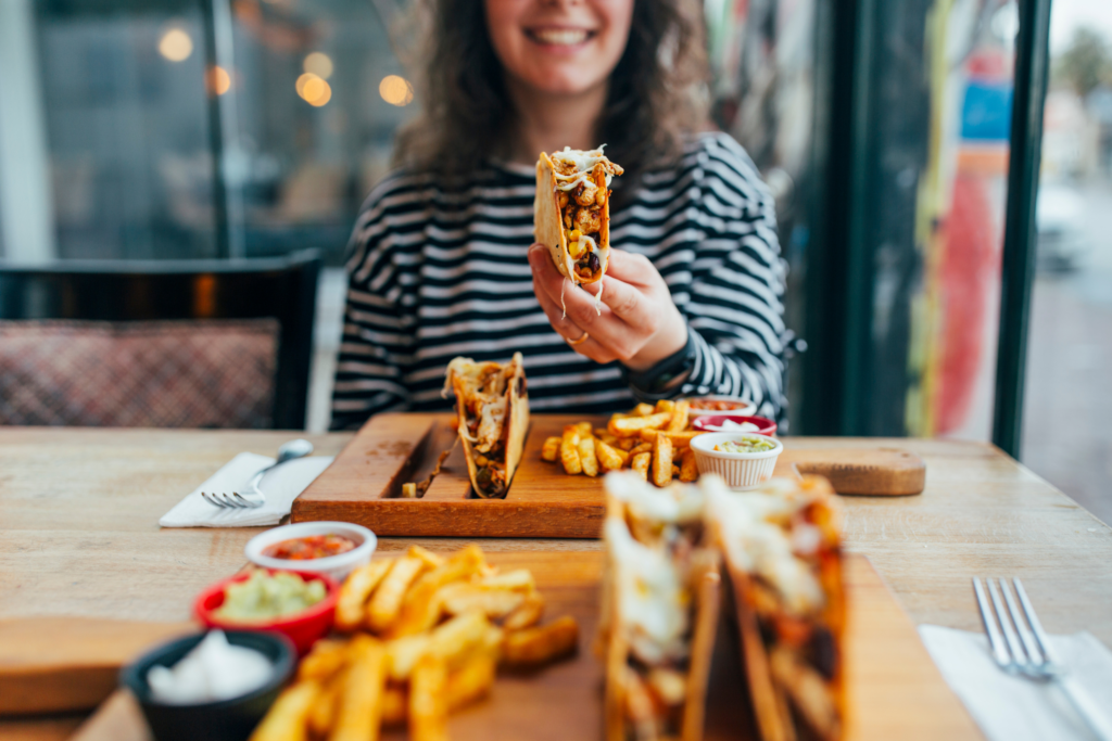 Picture is taken as if you are sitting across from a woman in a restaurant. In Front of you sits a wooden board with two hard shell tacos, fries, and taco dressings. Across from you the woman wears a black and white striped shirt and holds out a taco from her own board as she smiles.
