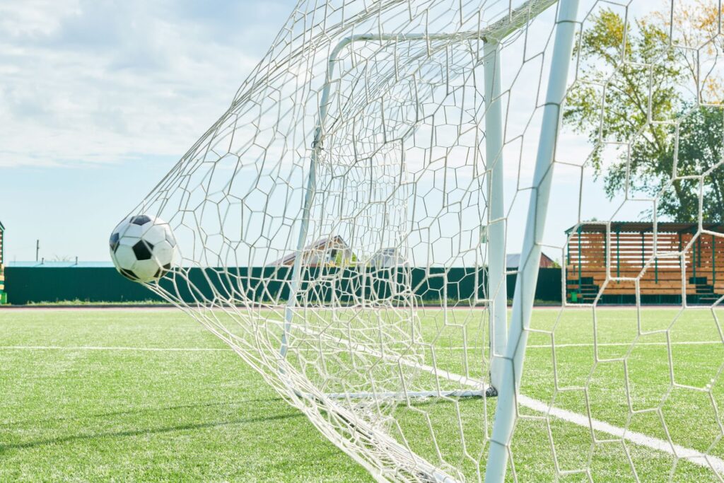 A soccer ball is hitting the back of a white soccer net on an outdoor soccer field. The net stretches with the ball. 
