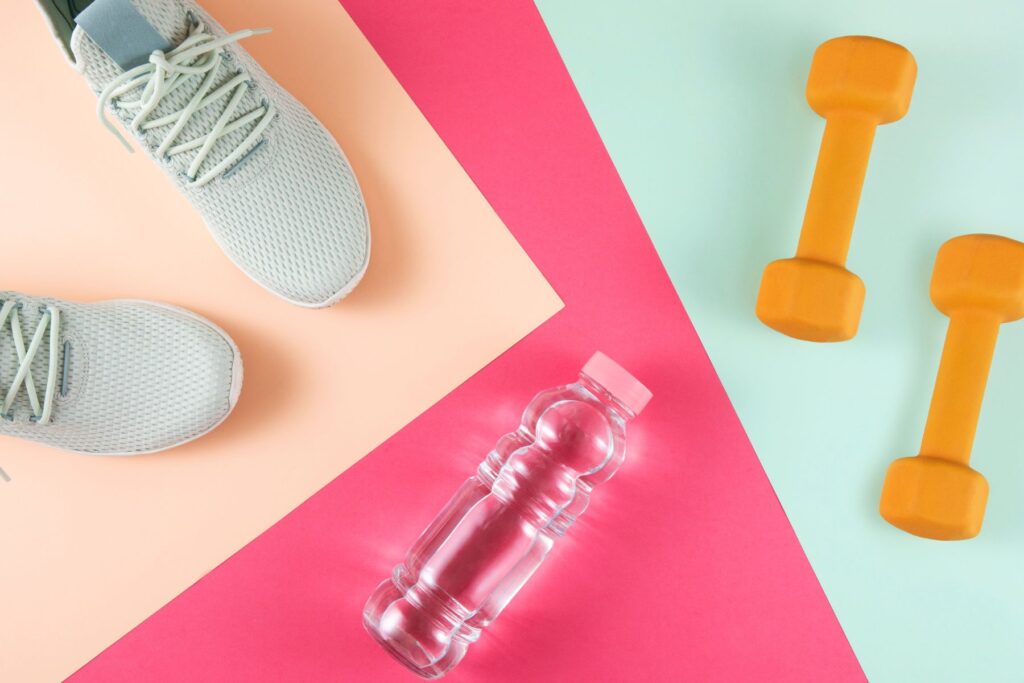 Overhead view of fitness related items including grey running shoes, water bottle, and orange dumbells on colourful background.