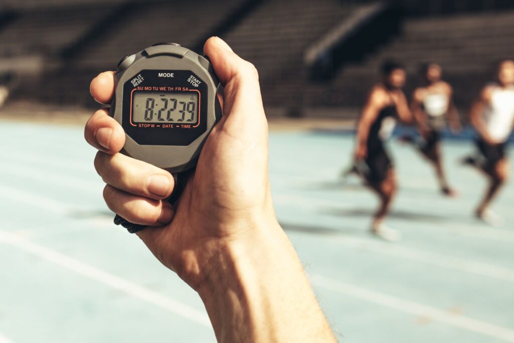 A man's hand holds up a stopwatch that reads 8 minutes and 22 seconds. In the blurred background, three athletes are running on a green track. An example of a different sports metric that does not involve weight.