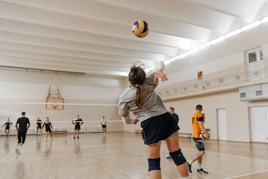 Woman jumping up to hit a volleyball over a net in a white gym. In the gym are other individuals also playing volleyball.