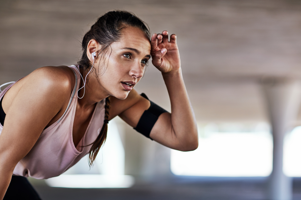 Female runner leaning over with her hand on her thigh as she wipes her brown with her other hand as if she is sweating and out of breath. THe runner has on a pick tank top. black arm band, and white earbuds. 