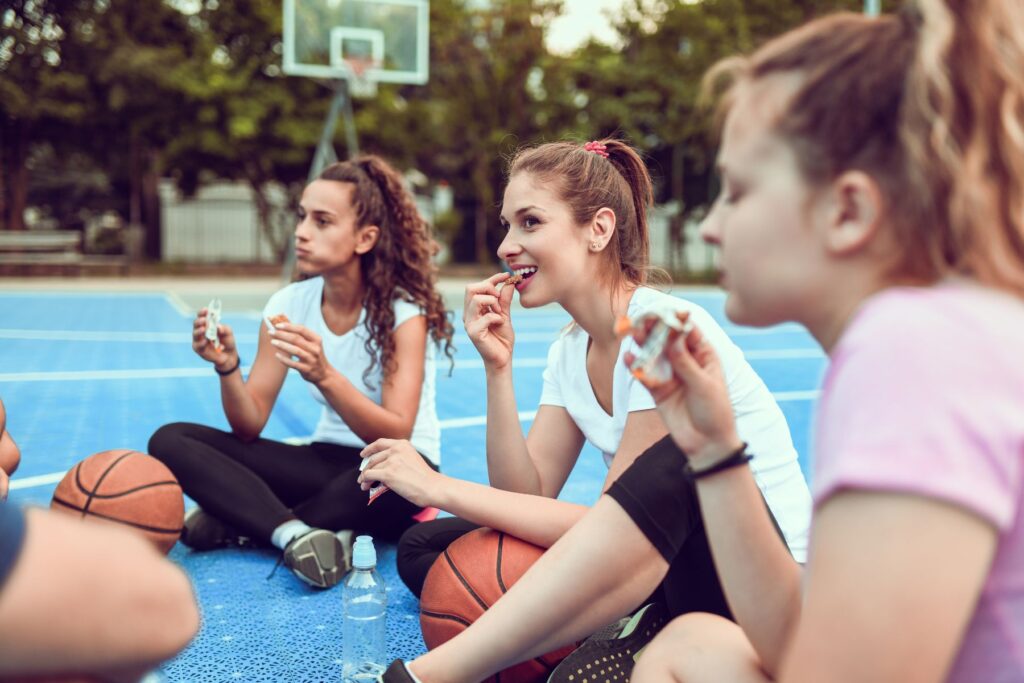 Three female athlete basketball players sit cross-legged together on the ground of an outdoor basketball court. The girls smile as they eat protein bars. 