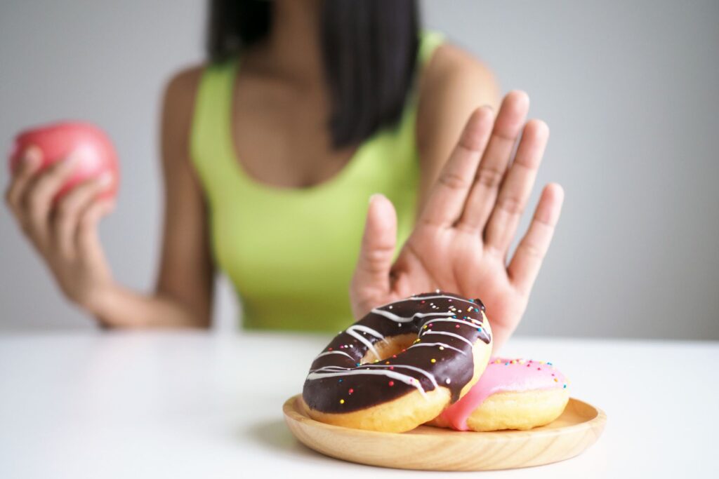 Image of female in neon yellow shirt pushing away doughnuts with one hand while holding an apple in the other as if she is refusing to eat the doughnuts.