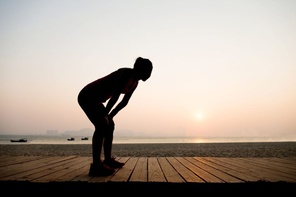 The silhouette of a woman who appears to be running on a boardwalk at a beach at sunset. The woman is bent over with her hands on her knees as if she is tired and catching her breath. 