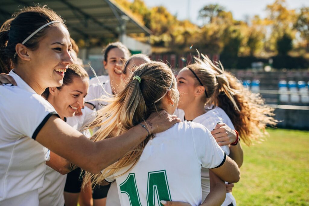 A group of female soccer players smile as they huddle together. The girls have their hair tied in pony tails and wear white and green soccer uniforms. THey appear to be outside on a grass field. 