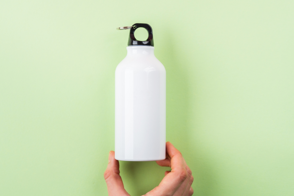 A white reusable water bottle with a black lid that can be used for staying hydrated is held up by a hand against a green background. 