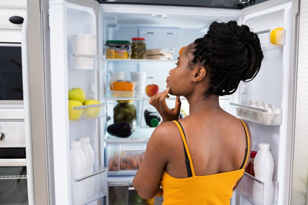 A woman looking into her fridge that is filled with food as if she is thinking about what to choose to eat. The image is taken as if the viewer is standing right behind the woman.