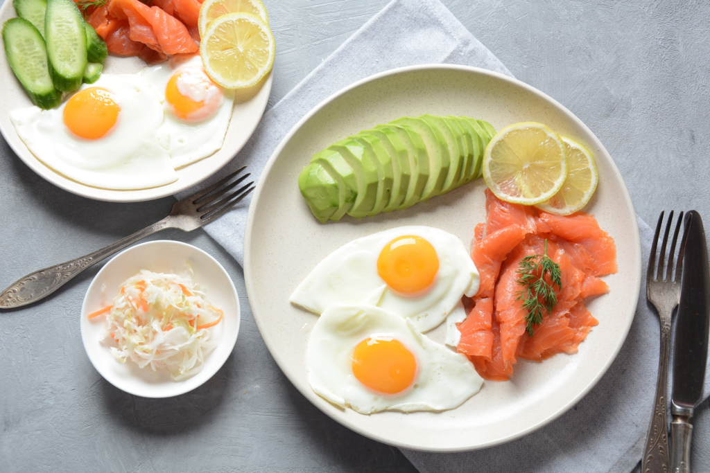 2 white plates with 2 fried eggs, smoked salmon, sliced avocado, and lemon slices sit on a grey countertop. A small white bowl with coleslaw and silverware sit beside the plates. 