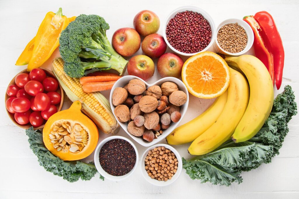 Overhead view of a spread of high fiber fruits, vegetables, nuts, and grains including banana, corn, chestnuts, kale, tomatoes, broccoli, red quinoa and more. 