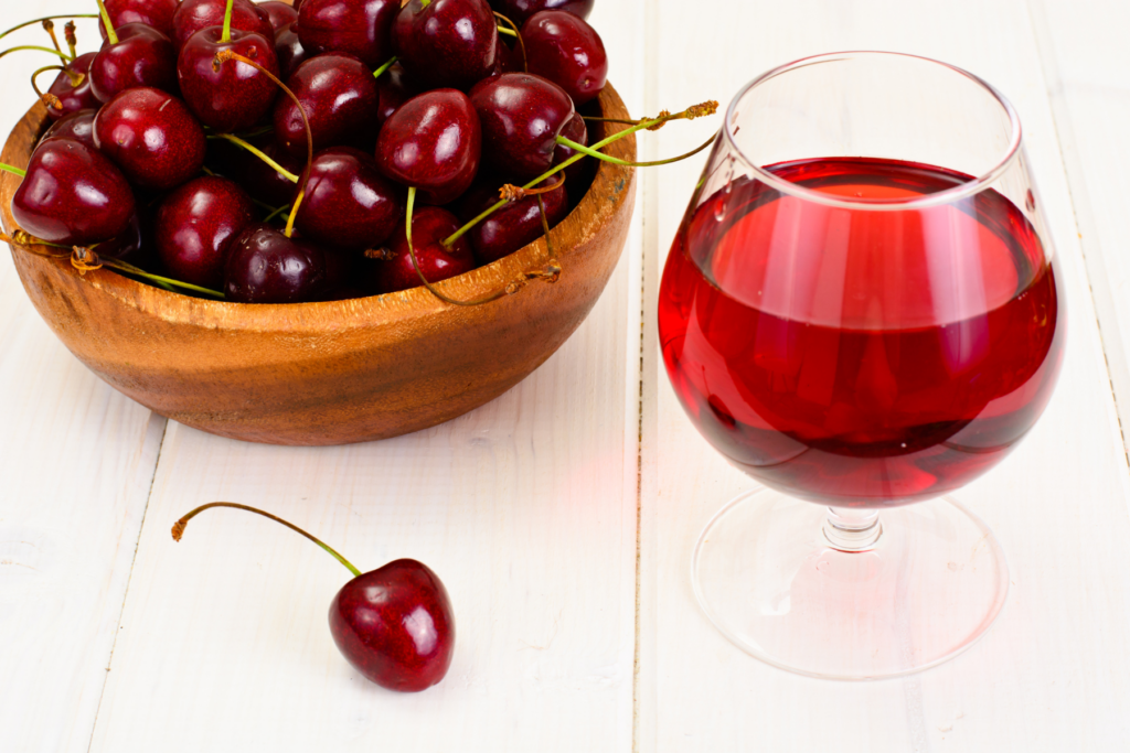 On the right side sits a wine glass filled with red tart cherry juice. On the left sits a wooden bowl filled with red cherries. The backdrop is white. 